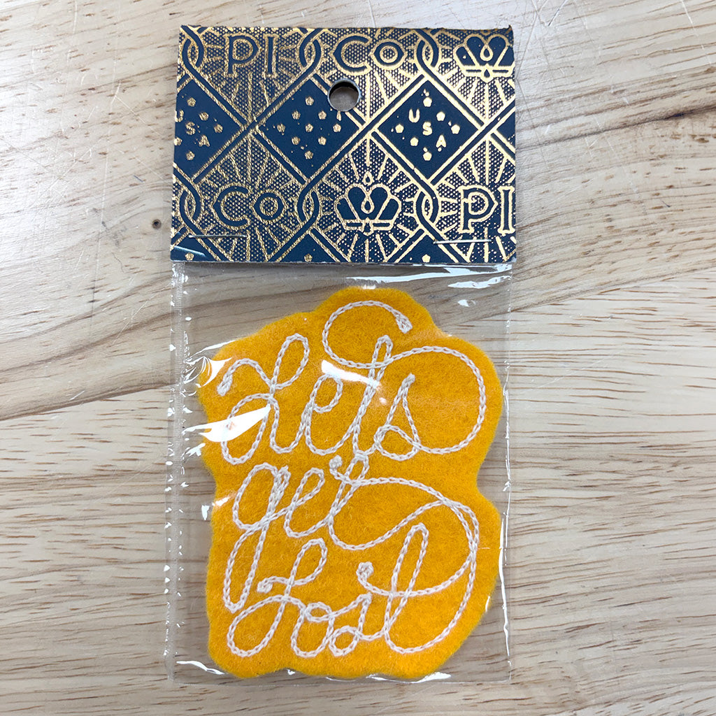 A yellow felt patch with white embroidered cursive text reading 'Lets Get Lost' inside a clear plastic bag, featuring a navy blue and gold patterned header card, placed on a light wood surface.