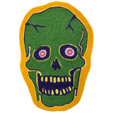 A green skull-shaped patch with a yellow border, featuring dark purple facial details and pink-and-white eyes, shown on a plain white background.