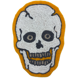 A skull-shaped chainstitch patch with a textured white skull design outlined in black, set against a mustard yellow background.