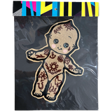 A decorative embroidered patch shaped like a baby figure, shown inside clear plastic packaging with a black backing and a colorful striped header card. The patch has a tan base with dark brown tattoo-style patterns and the word 'BABY' embroidered on the forehead.