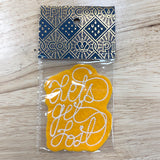A yellow felt patch with white embroidered cursive text reading 'Lets Get Lost' inside a clear plastic bag, featuring a navy blue and gold patterned header card, placed on a light wood surface.