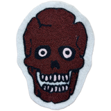 A handcrafted chainstitch patch featuring a stylized skull with a dark reddish-brown textured face, black outlines, and white details, set against a light gray background.