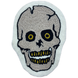 A skull-shaped embroidered patch with a textured beige skull design, black outlines, and yellow details on a white felt background.