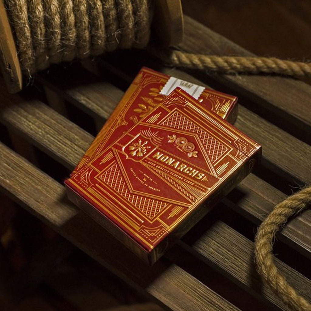 A red deck of Monarch playing cards with gold detailing, resting on a wooden surface with a rope in the background.